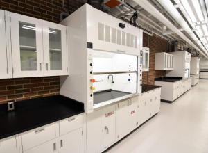 A newly constructed laboratory space with white cabinets, a large white fume hood, and black countertops. The brick wall behind the workbenches contrasts with the bright, open design, and overhead fluorescent lighting illuminates the spacious hallway layout.