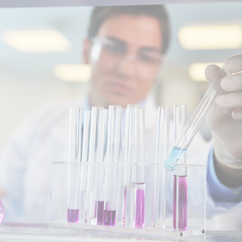 Male scientist with short hair and wearing safety glasses working in a laboratory. In the foreground there is a set of test tubes held in a stand, some containing a purple liquid and one being filled with a pipette that has a blue liquid.