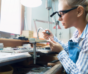 A jewelry artisan wearing magnifying glasses and a blue apron is meticulously working on a small piece of jewelry at a well-lit workbench.