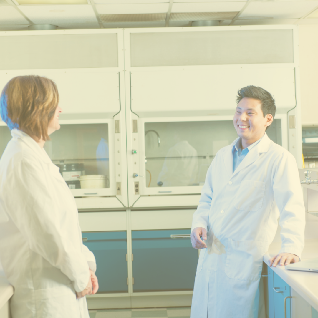 overexposed photo of two people in a laboratory environment. Both individuals are wearing white lab coats, as they are scientists or researchers. The person on the left is a woman with short hair, facing the man on the right, who is smiling. They are having a conversation. In the background, there are fume hoods with closed sashes.
