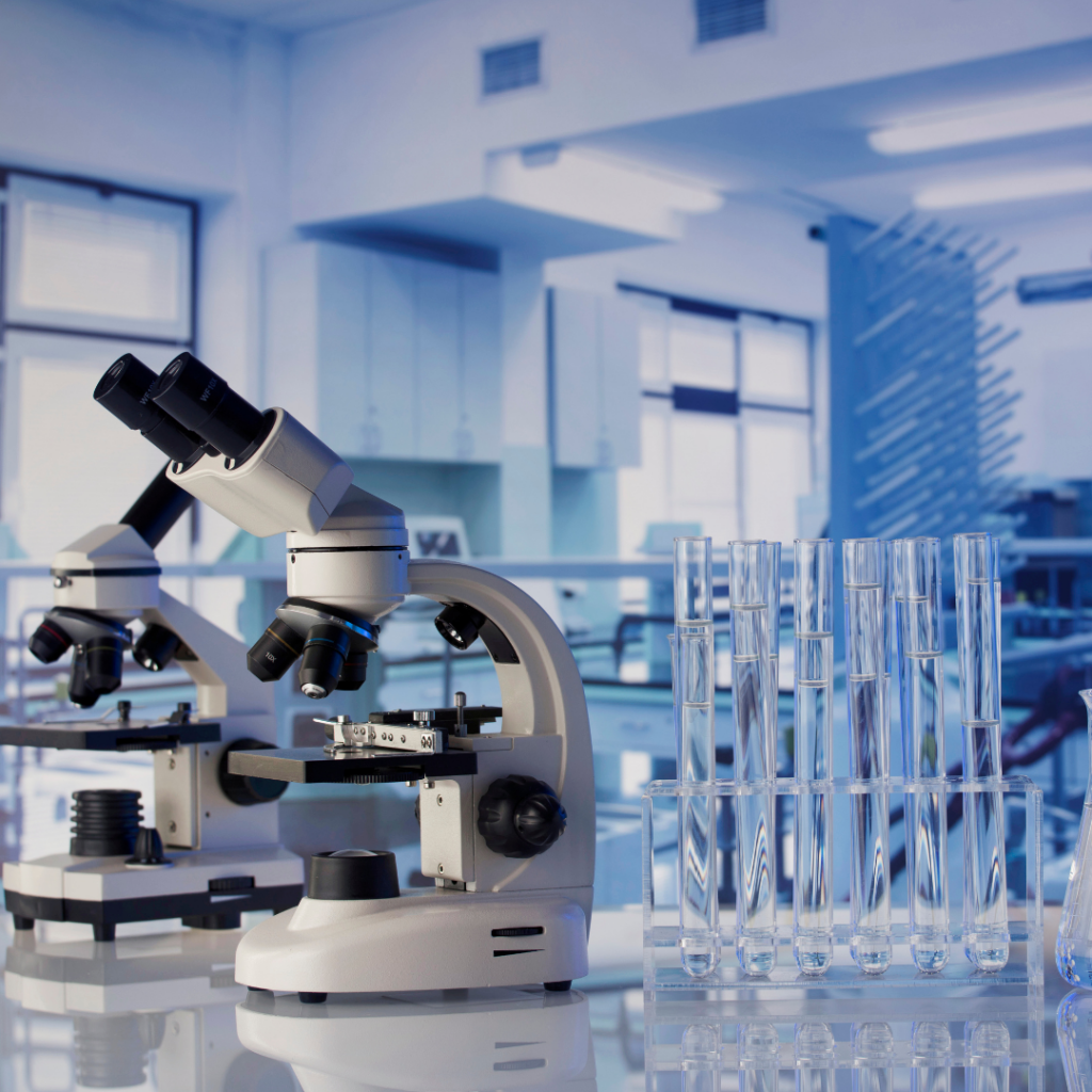 Two light-colored microscopes are positioned on a glossy lab bench alongside a clear rack holding multiple glass test tubes. The background reveals a spacious, modern laboratory with cabinets and bright windows, creating a clean, scientific atmosphere.