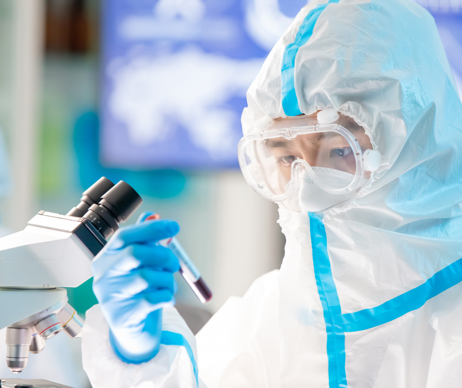 A laboratory technician wearing full personal protective equipment (PPE), including a white protective suit with blue seams, goggles, a face mask, and blue gloves, examines a test tube containing a dark red liquid.