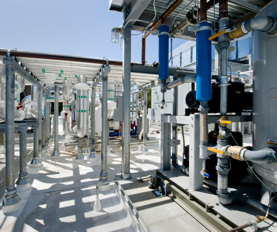 An outdoor industrial installation filled with interconnected metal pipes, cylindrical components, and valves under a partial roof structure. Several pipes are wrapped in insulation, while others feature blue filter housings and green labeling. Sunlight casts sharp shadows on the white surface as various machinery and equipment are arranged in a compact layout.