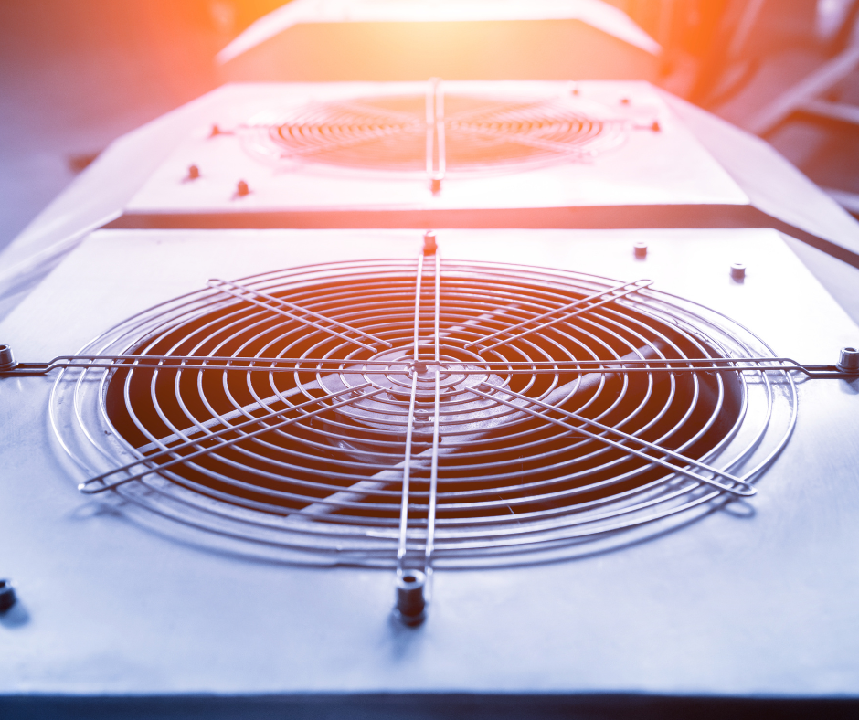 A close-up view of two large, circular metal fan grilles on top of industrial cooling units, illuminated by warm overhead lighting. The fan blades and protective wire mesh are clearly visible, emphasizing the equipment’s function in temperature regulation or ventilation.