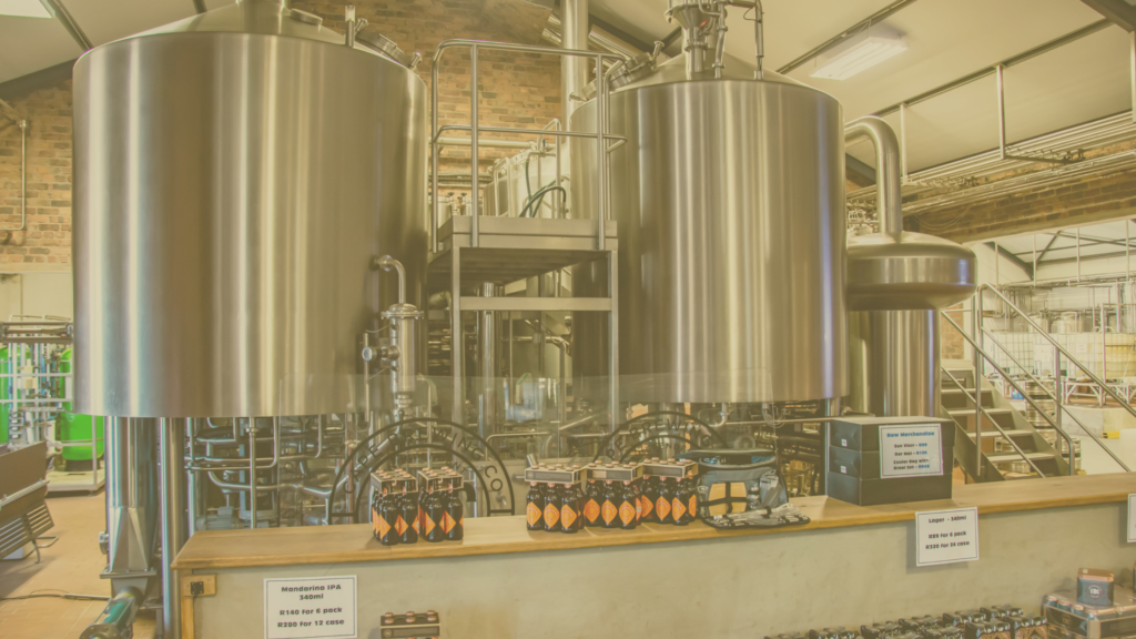 A brewery facility with large stainless steel fermentation tanks connected by pipes, valves, and platforms. The tanks are polished and cylindrical, reflecting the industrial environment. In the foreground, a wooden counter displays bottled craft beer with orange and black labels, neatly arranged in six-packs. Small signs with pricing information are placed on the counter, indicating different beer options.