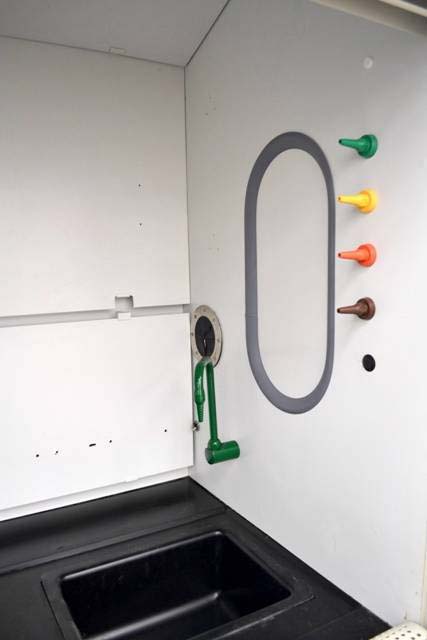 The interior of a laboratory fume hood, featuring a black work surface with a built-in rectangular sink. On the right wall, there are four color-coded control knobs (green, yellow, red, and brown) for utilities such as gas, air, and vacuum.