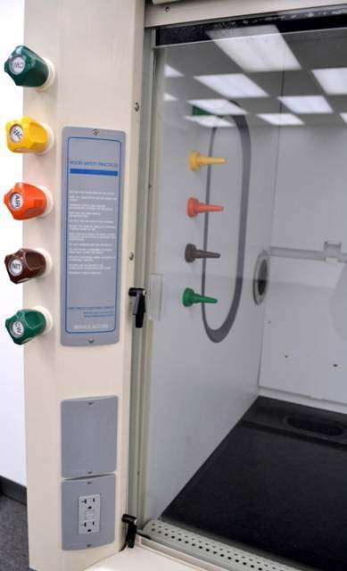 A laboratory fume hood with a transparent glass sash, showing the interior workspace designed for handling hazardous substances. On the left side of the fume hood, there are color-coded control knobs for different utilities such as gas, air, and vacuum, labeled with abbreviations like "AIR" and "NIT."