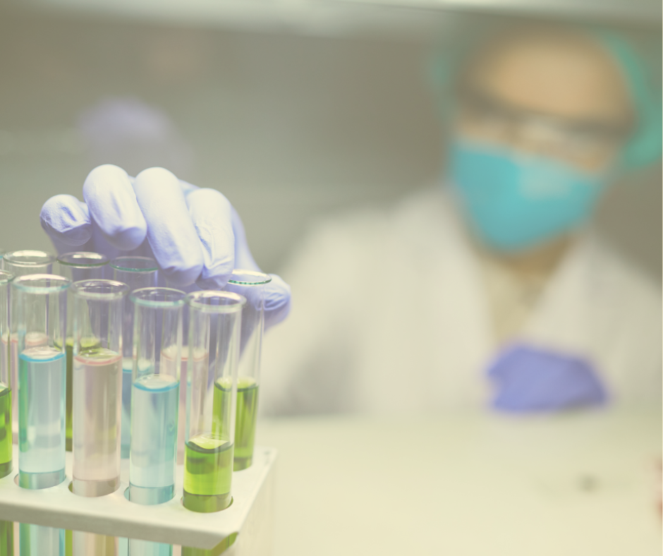 A laboratory technician wearing a lab coat, safety goggles, face mask, and purple gloves selects a test tube filled with green liquid from a rack of multicolored liquid-filled test tubes inside a fume hood or controlled environment.