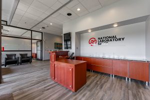 Modern office reception area for National Laboratory Sales, featuring a curved wooden reception desk with a glass countertop, wood-look tile flooring, and a wall-mounted company logo with a red circular emblem.